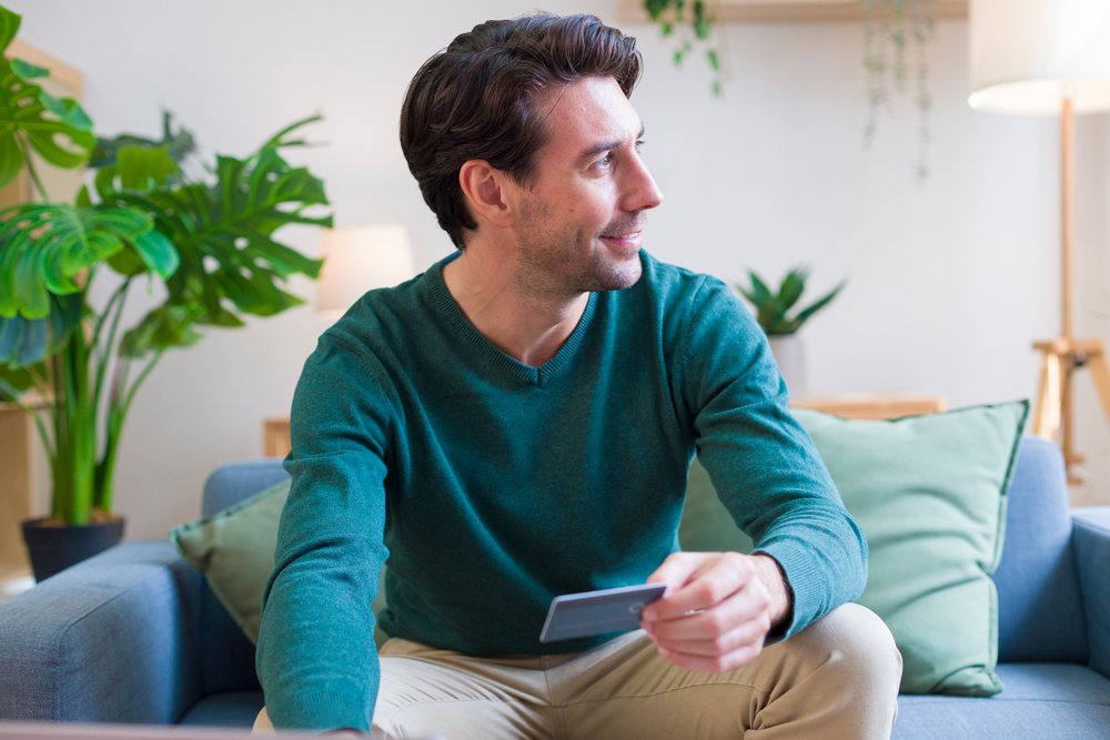 Man sitting on a sofa a credit card and smiling in a modern living room, technology, young adult, home.