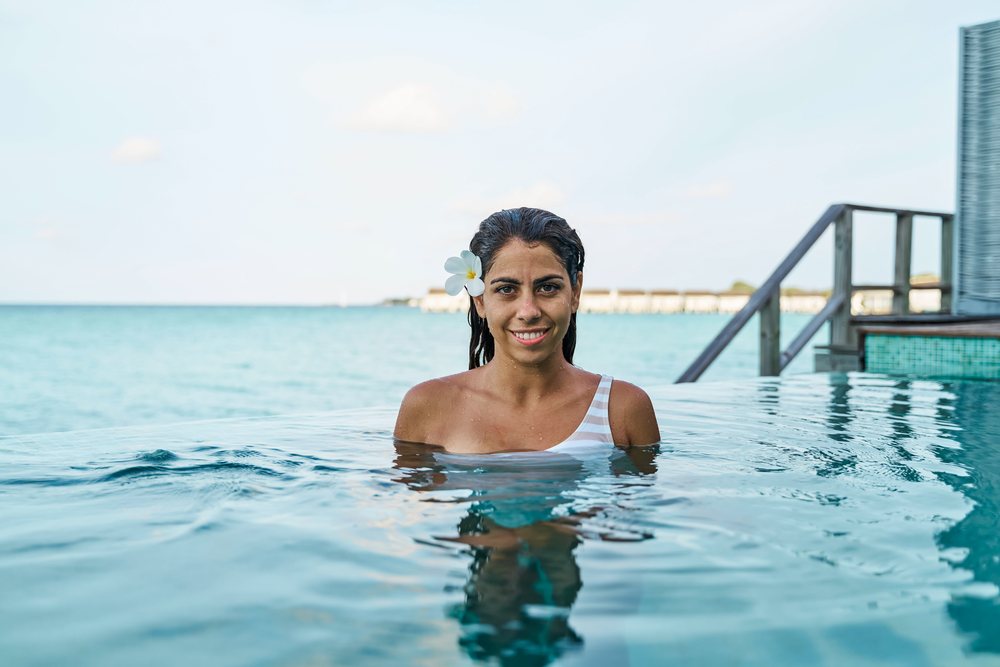 A smiling white woman enjoys an infinity pool with ocean view