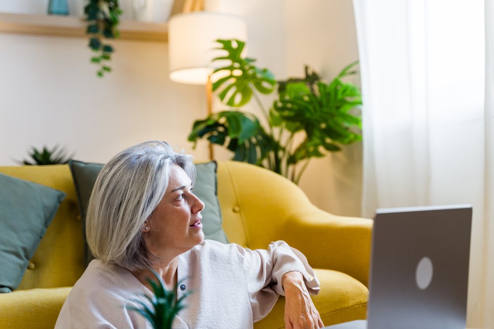 Woman with gray hair sitting on the floor next to a yellow sofa and laptop in a bright living room decorated with plants, Spain.