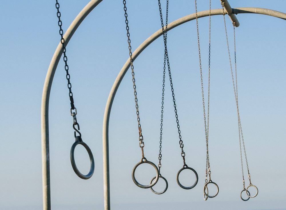 Multiple gymnastic rings hang from metal chains attached to metal arch frames outdoors under a clear blue sky, California, USA.