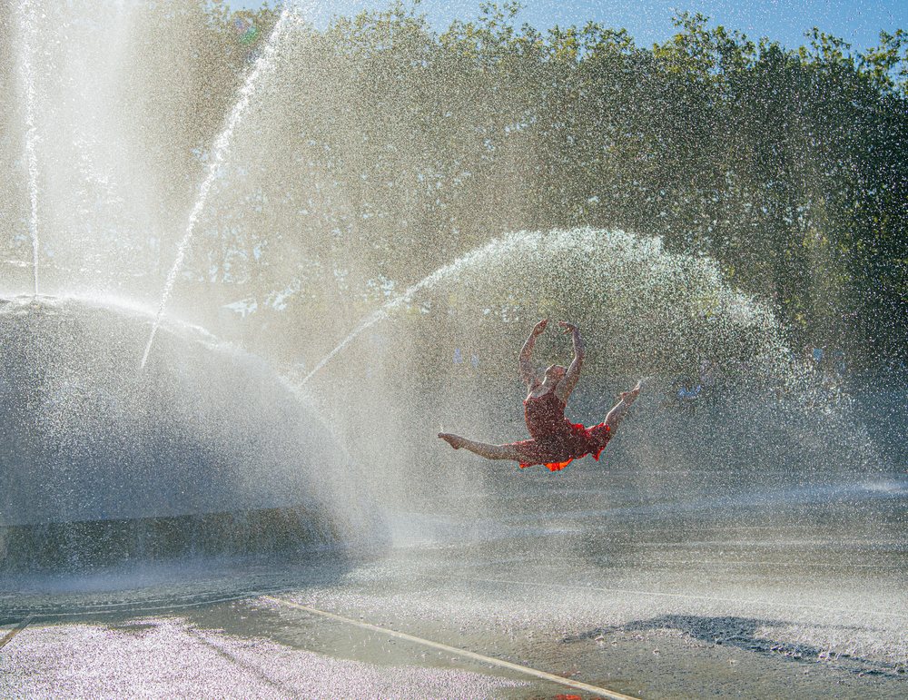 Dancer in a red dress leaps mid-air through water sprays from a fountain in an outdoor park, USA.