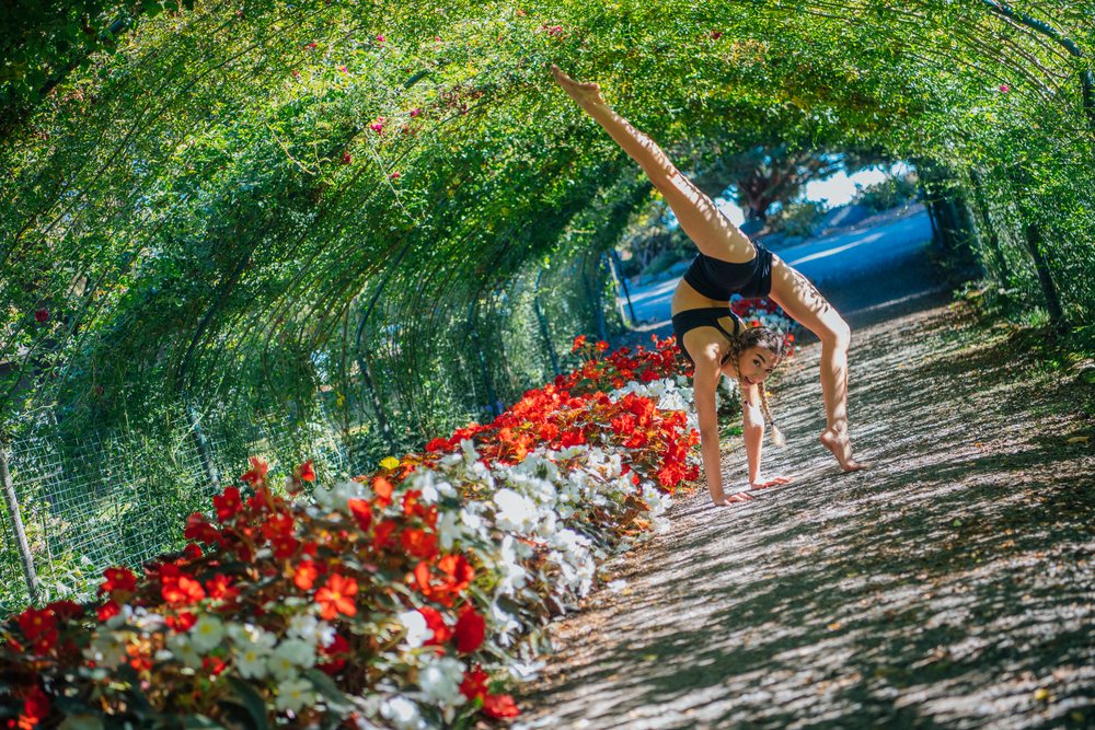 Woman in black sportswear performs a contortionist handstand pose inside a flower-lined garden tunnel, Washington, Tacoma, USA.