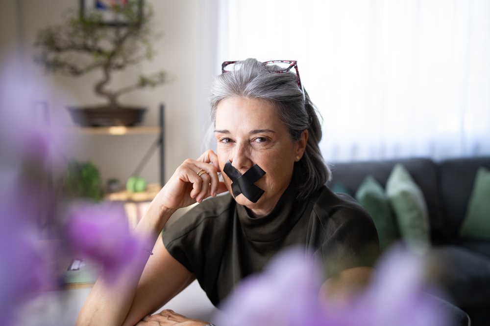 Gray-haired woman sits indoors in a living room with black tape over her mouth, wearing glasses on her head and a thoughtful expression, Spain.