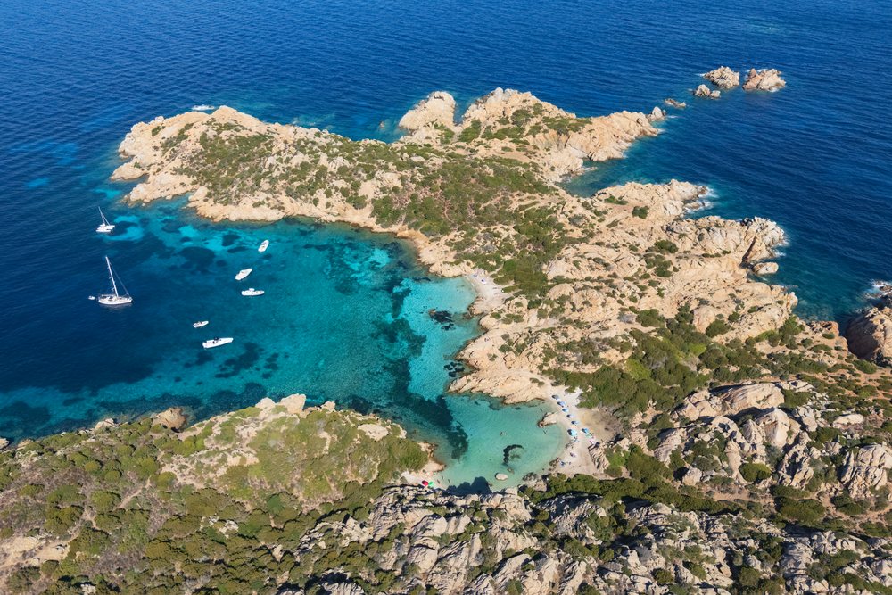 Cala Napolatana beach and cove feature clear turquoise water, rocky coastline with shrubs, and boats anchored near the sandy shore, Travel Images, La Maddalena, Sardinia, Italy.