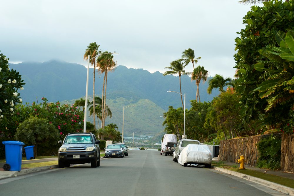 Asphalt street in a residential neighborhood, lined with palm trees, tropical plants, parked cars, and blue recycling bins under cloudy skies, Oahu, Hawaii, USA.