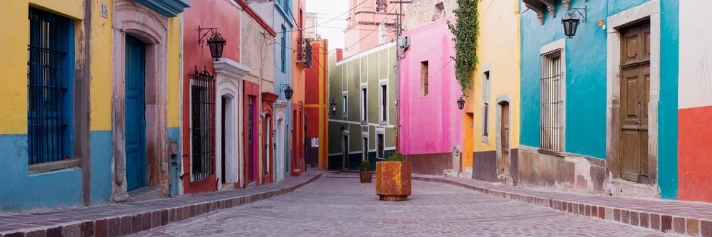 Colorful colonial-style buildings line a narrow cobblestone pedestrian street, decorated with ivy and plant pots, Guanajuato, Mexico.