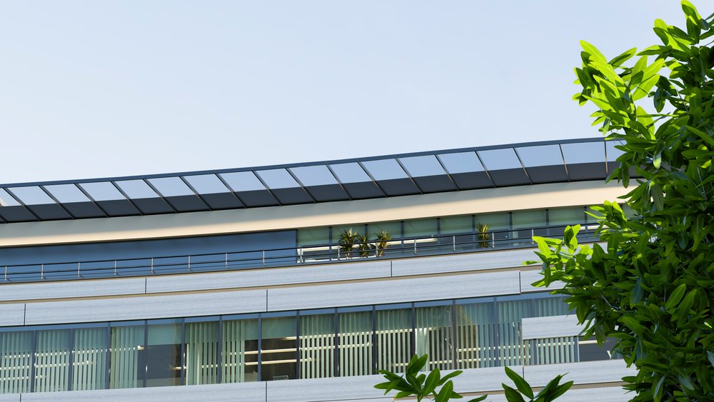 Modern office building exterior featuring glass windows, balcony with potted plants, and green leaves against clear blue sky.