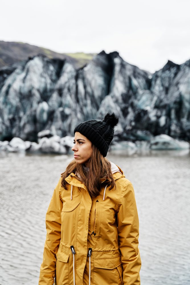White Woman in yellow jacket standing near glacier water in Iceland