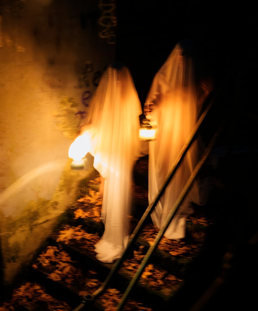 Two ghost figures holding lanterns descend outdoor stairs covered with autumn leaves at night, ghosts, USA.