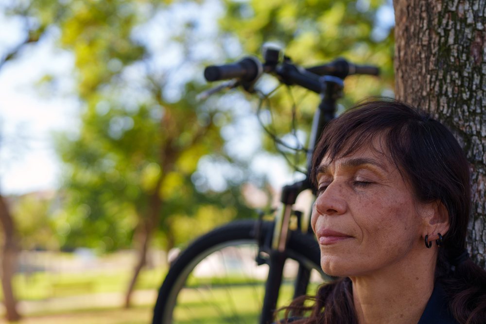 Woman with closed eyes relaxes leaning against a tree in an outdoor park with a bicycle behind her, Málaga.