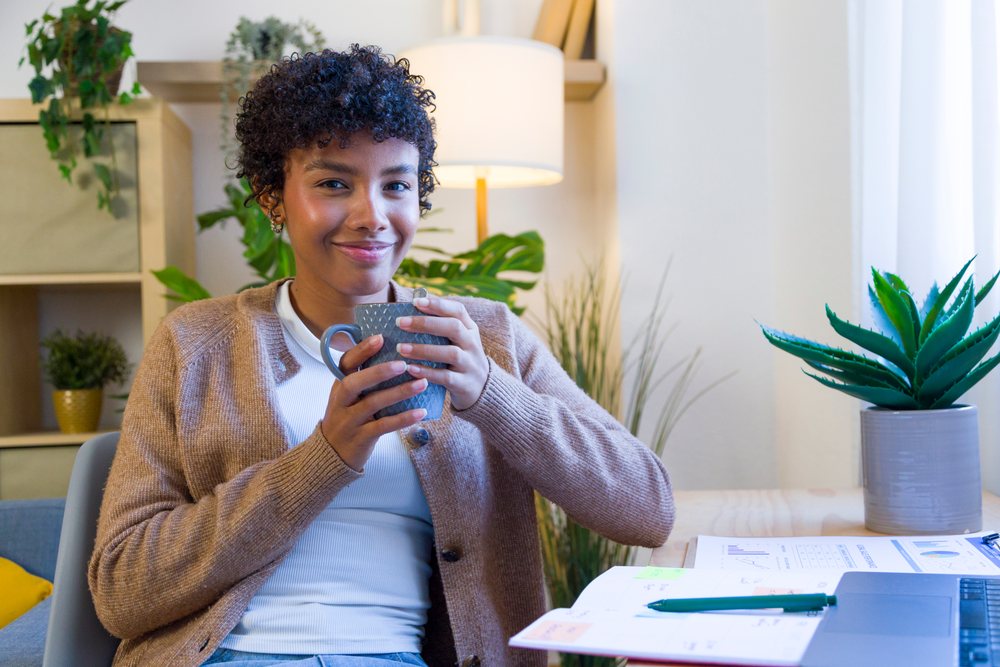 Young woman holding a mug and smiling while seated at a desk with papers and plants in a cozy home office.