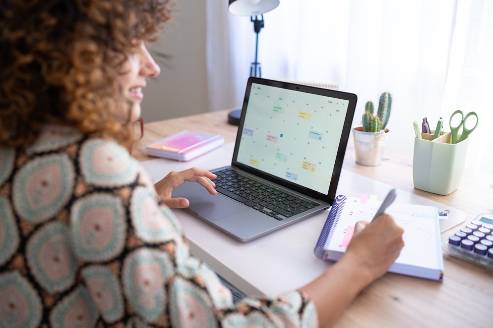 Woman writes in a spiral notebook while viewing a monthly calendar on a laptop at a home office desk with plants and office supplies, Spain.