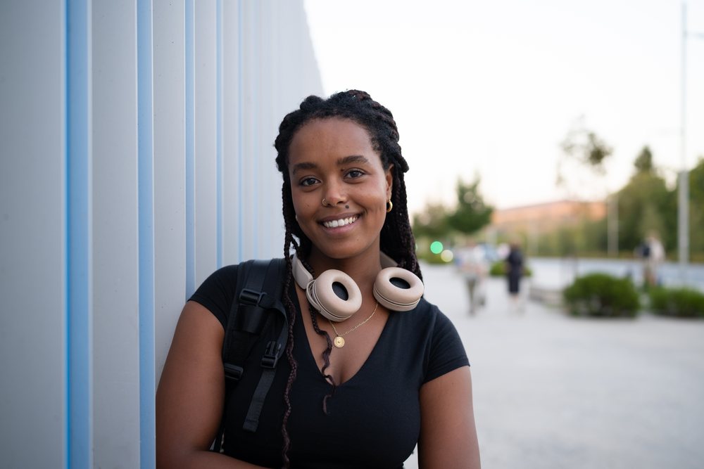 A young woman with braided hair and headphones around her neck smiles while leaning against a metal wall in an urban setting, Santiago De Compostela, Spain.