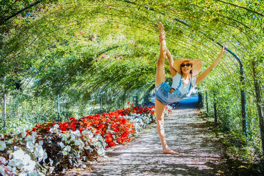 Barefoot young woman wearing denim overalls and a sun hat performs a contortion stretching pose in a flower-covered garden tunnel, contortionist, Tacoma, WA, USA.