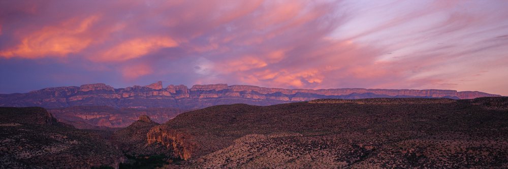Sunset sky over rugged mountain range and arid desert terrain with colorful purple and orange clouds in clear sky.