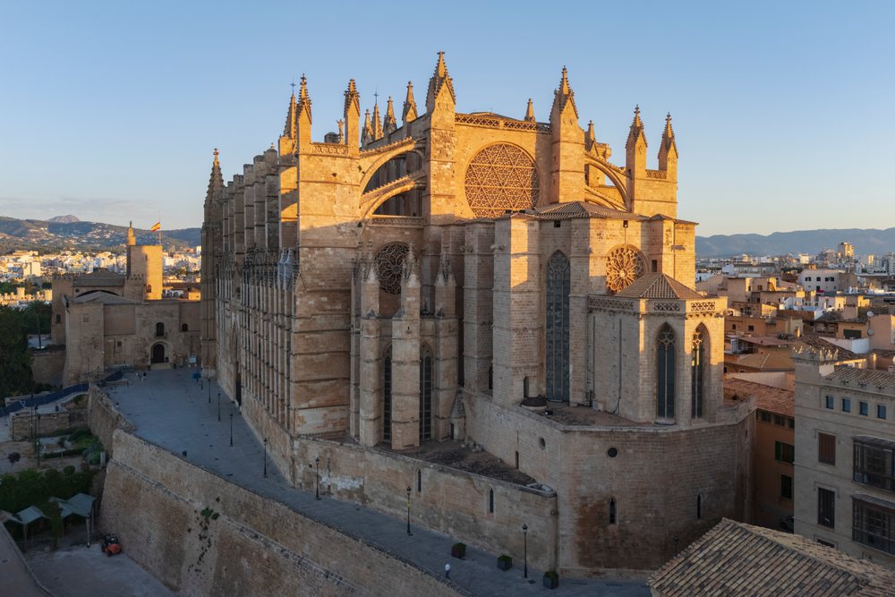 Gothic Cathedral with stone masonry, rose windows, surrounding cityscape, fortification walls, paved walkways, and mountains in the background at sunset, Landscape, Lifestyle, Palma de Mallorca, Balearic Islands, Spain.