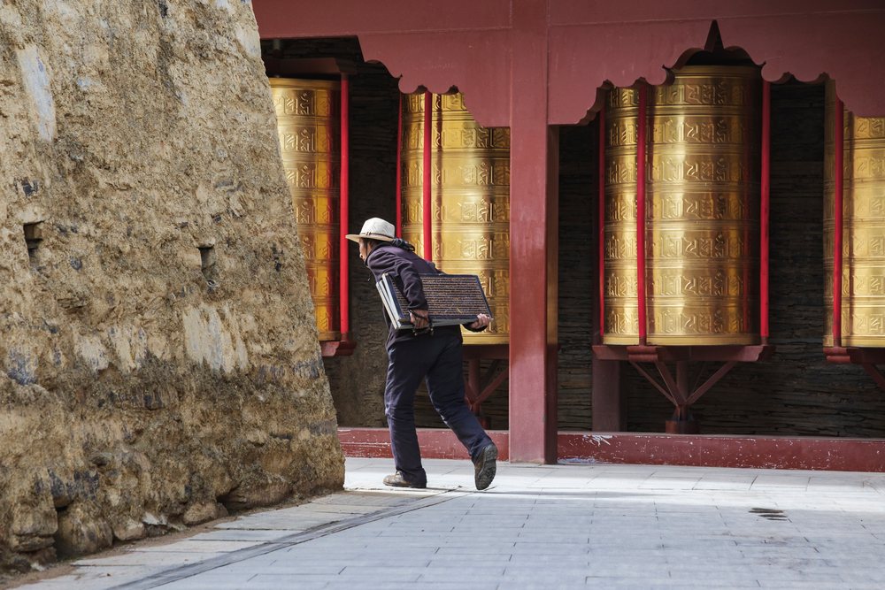 Tibetan man wearing a hat walks along a paved path carrying an inscribed wooden tablet toward a Mani Wall with large golden prayer wheels, Tagong, Sichuan Province, People's Republic of China.