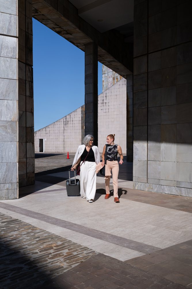 Two businesswomen walk and talk beside modern architectural columns outdoors on a sunny day, Spain.