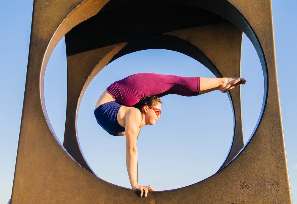 Woman performs a handstand yoga pose on a circular metal sculpture outdoors under a clear blue sky, USA.