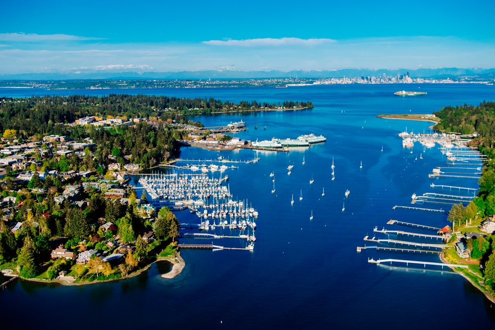 Aerial view of Bainbridge Island's Eagle Harbor marina with boats and piers, overlooking Puget Sound and the Seattle skyline in the distance, Salish Sea, USA.
