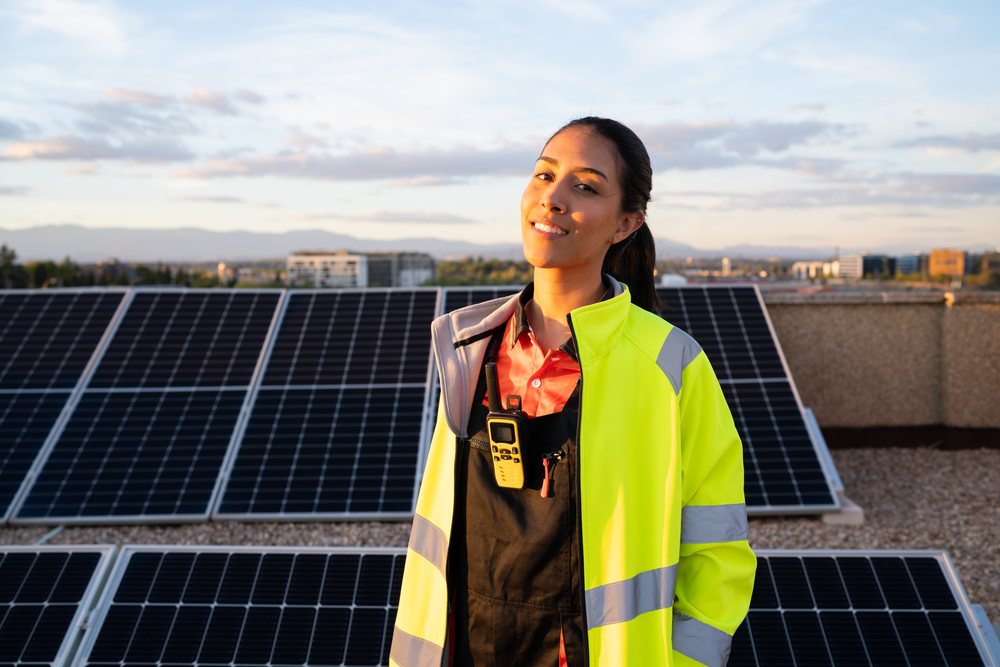 Woman engineer in high visibility jacket holding a walkie talkie on a rooftop with solar panels at sunset and mountains beyond a cityscape, Madrid, Spain.