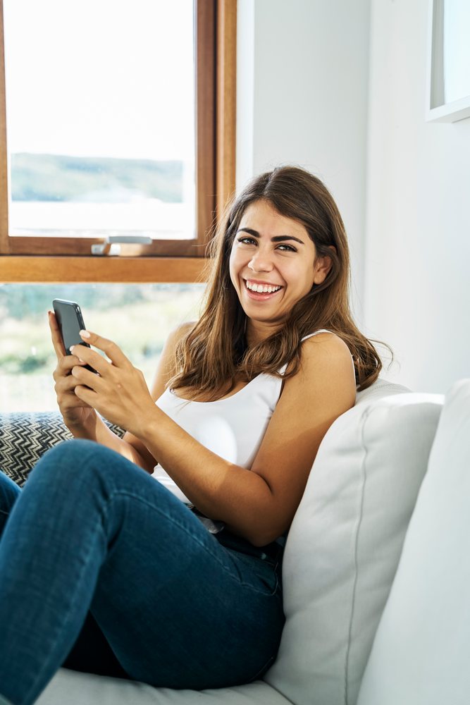A smiling young white woman sits on a couch using a smartphone