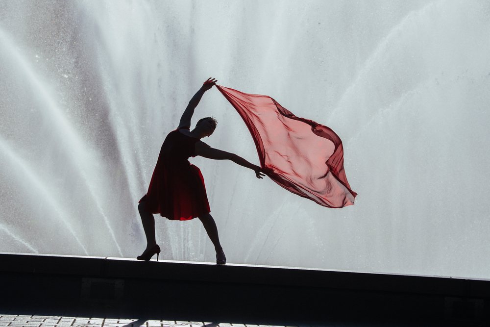 Silhouette of an adult woman dancer wearing a red dress and high heels, holding a flowing red scarf by a water fountain outdoors, USA.