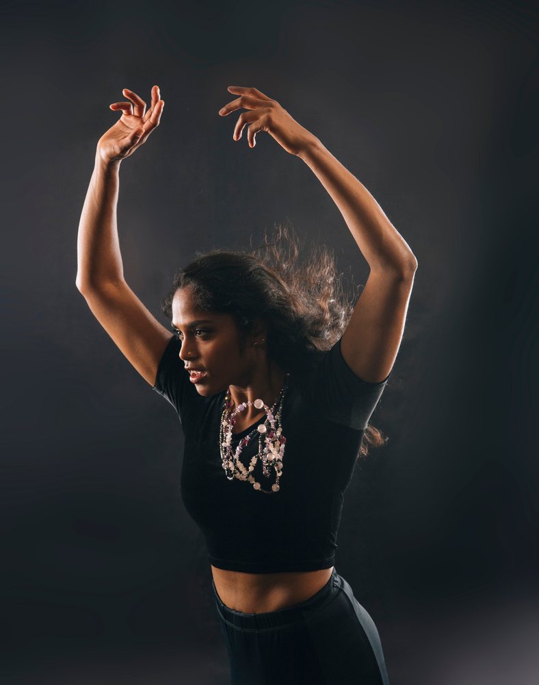 Woman dancing with raised arms wearing a black crop top and layered necklace in a dark studio background.