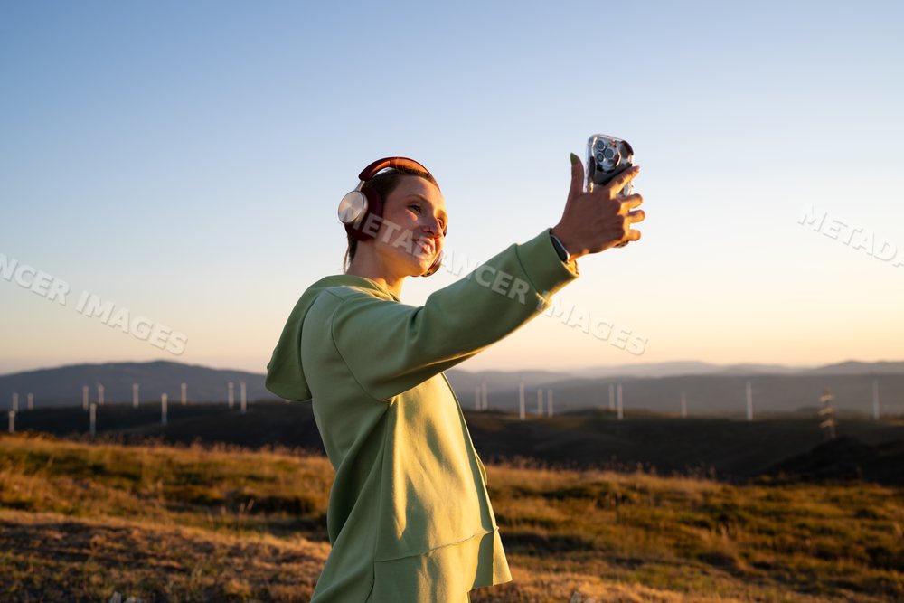 Young white woman takes a selfie outdoors near wind turbines at sunset