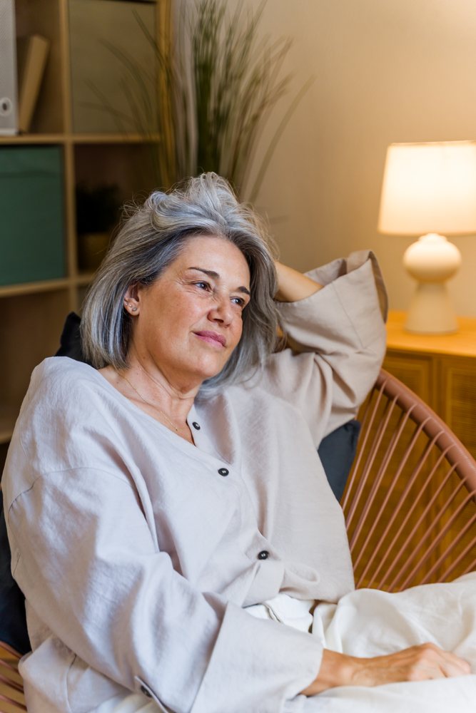 Mature woman with gray hair relaxing in an armchair in a contemporary indoor setting with a lamp and plants, Spain.