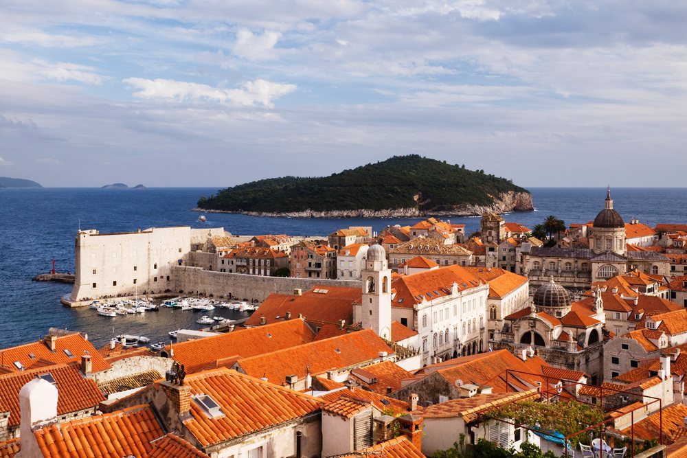 Historic old city of Dubrovnik with stone walls and red tile roofs beside green tree-covered Lokrum Island on the Adriatic Sea.