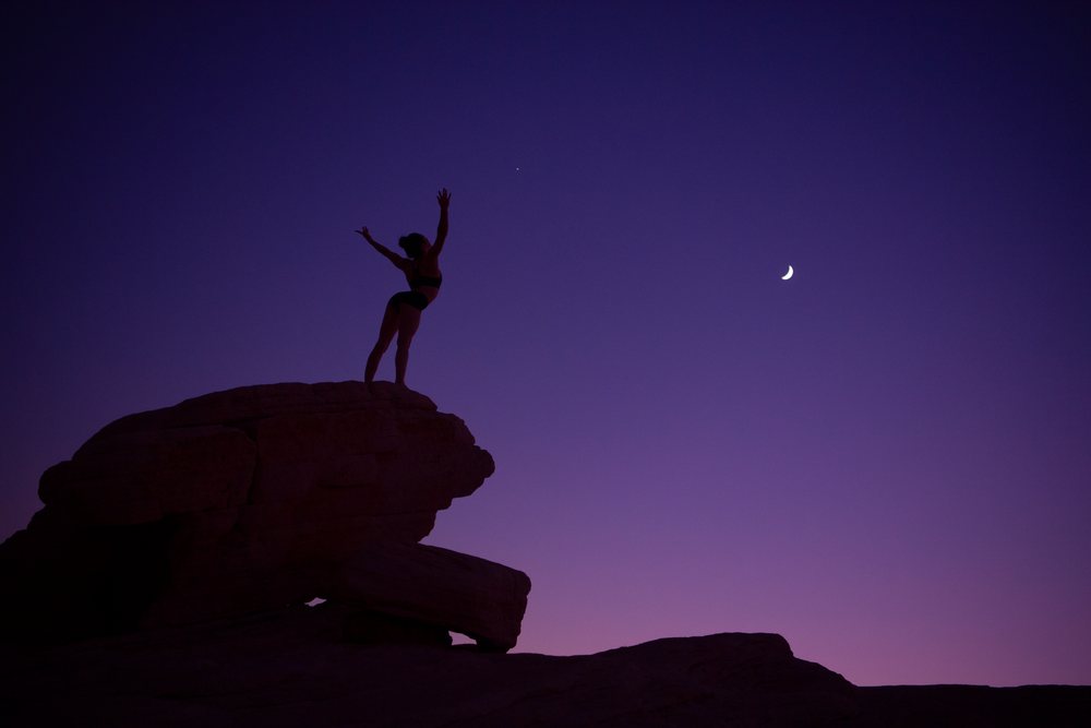Woman silhouette practicing yoga on a rock at dusk with a crescent moon in a natural outdoor setting, USA.
