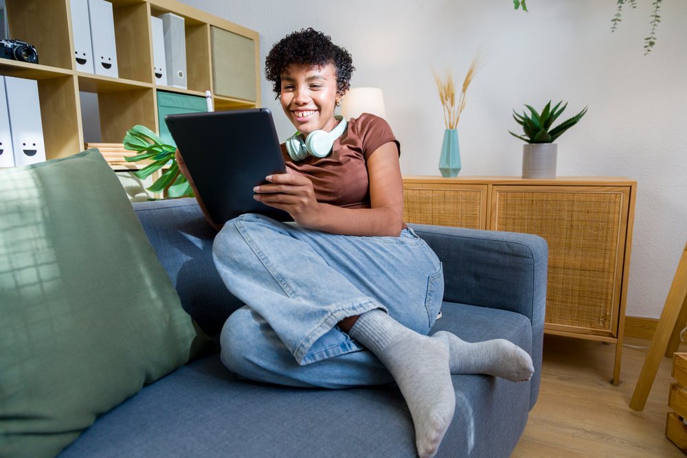 Young woman wearing casual clothing sits on a couch smiling while holding a tablet in a cozy living room with wooden furniture and indoor plants.