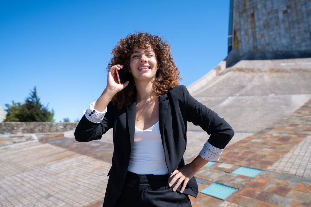 Businesswoman talking on phone outdoors by modern stone architecture, Santiago de Compostela, under a clear blue sky, traveling executive, Spain, Santiago de Compostela.
