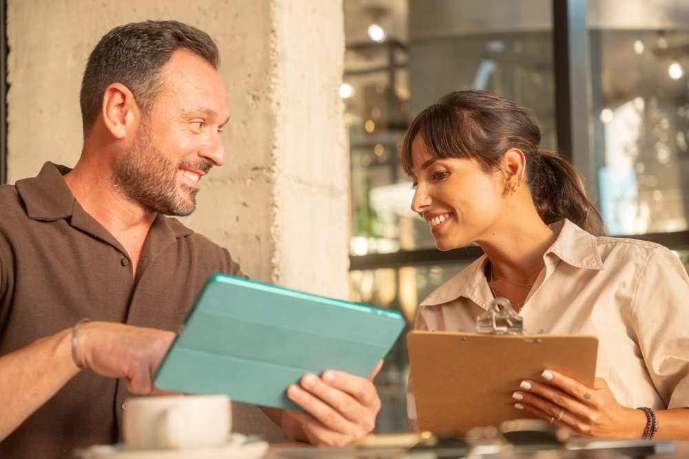 Two coworkers indoors, Andalucia share information using a tablet and clipboard during a business meeting, Seville.
