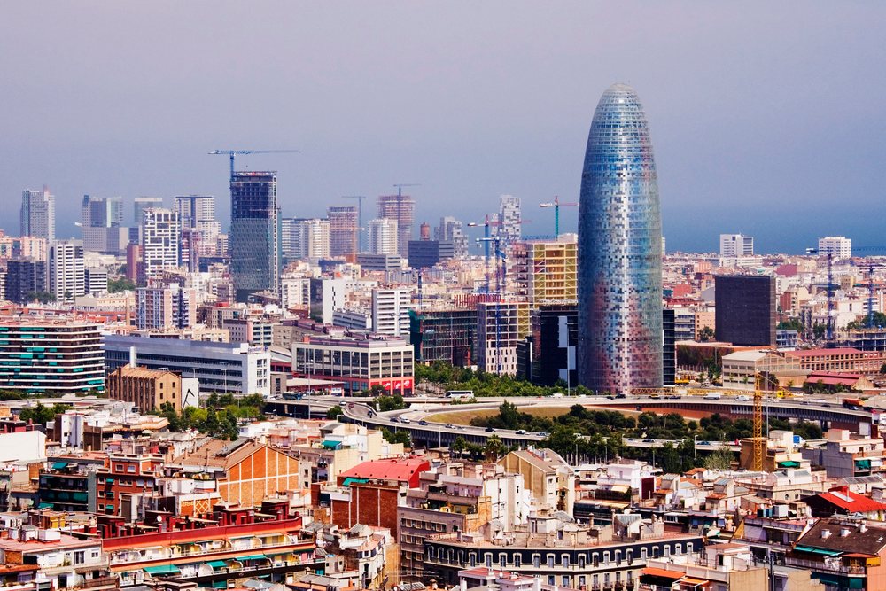 Barcelona cityscape showing Torre Glòries and Avenida Diagonal skyline on a clear summer day by the Mediterranean coast with visible construction cranes and traffic.