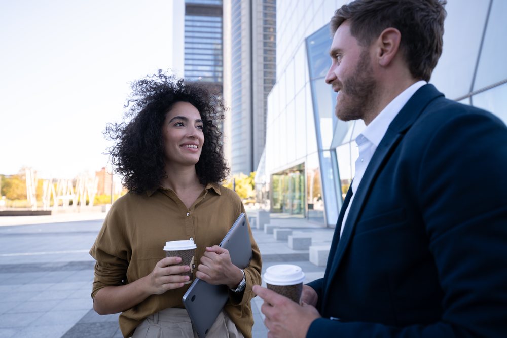 Two businesspeople hold coffee cups and a laptop while conversing outdoors near a modern office building, Madrid, Spain.