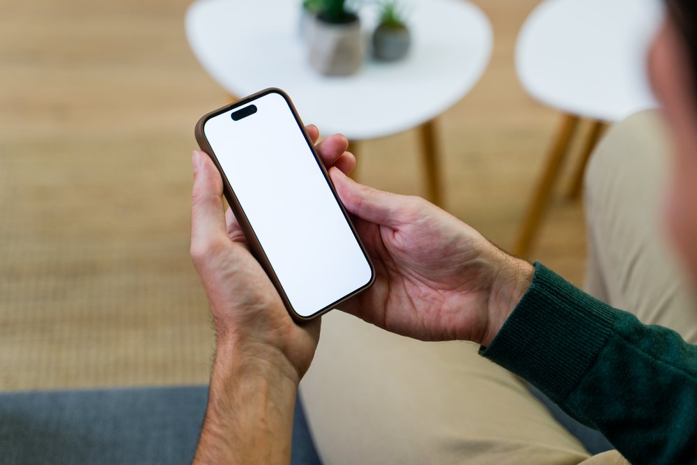 Man wearing green sweater holding smartphone with blank white screen near white table and wooden floor indoors, Spain.