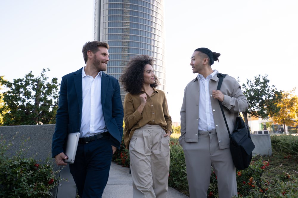 Three business professionals walking and conversing on an urban park sidewalk near a modern office building, Madrid, Spain.