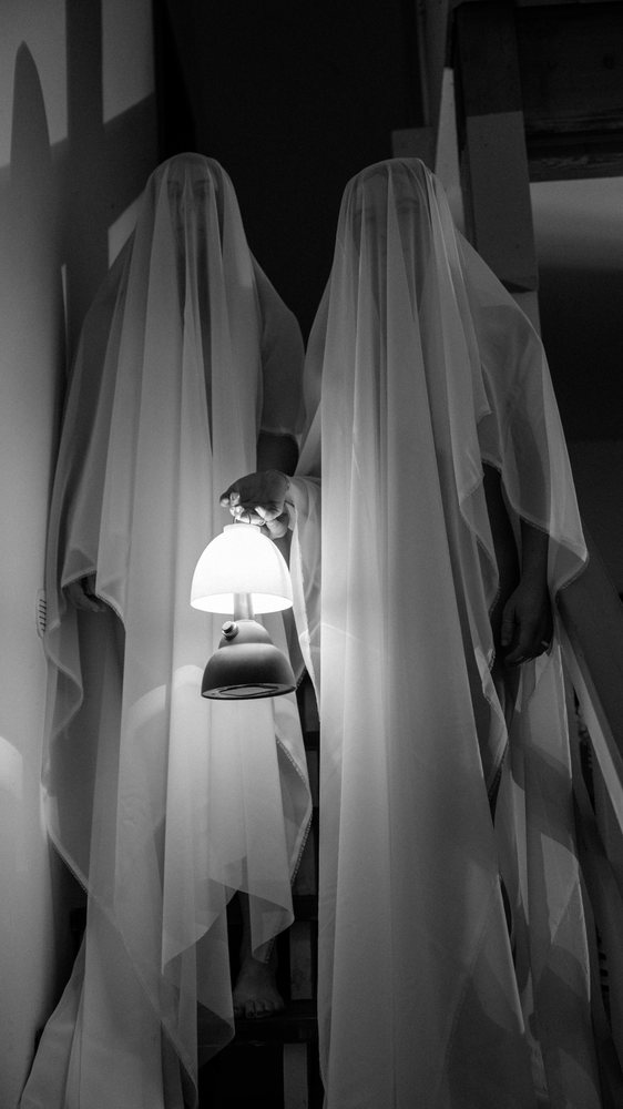 Two ghost figures stand barefoot on indoor stairs holding a lit lamp in a dimly lit indoor setting, ghosts, USA.
