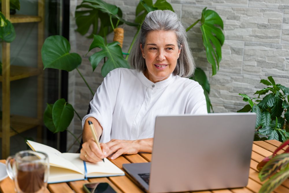 White Woman writing in notebook using laptop at wooden table