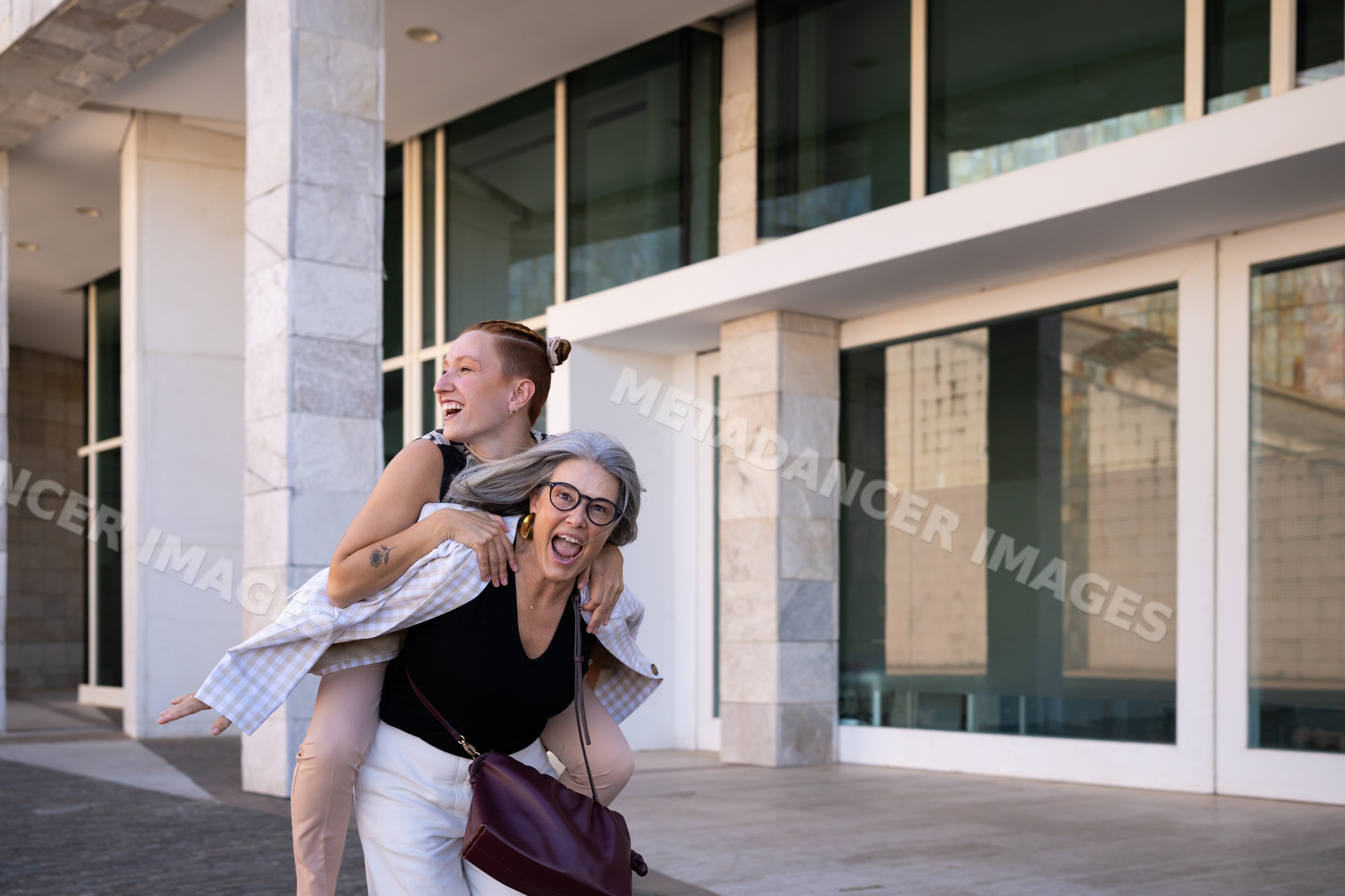 Two white women share joyful piggyback moment outside modern building