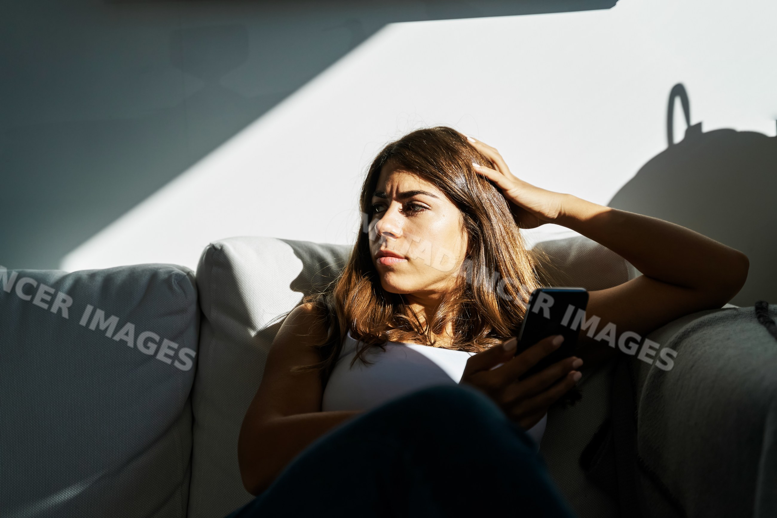 Young white woman holding mobile device sitting thoughtfully on couch
