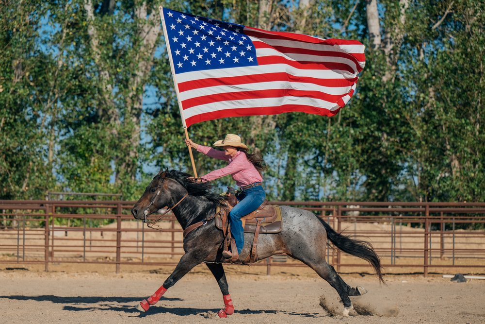 White woman riding horse carrying american flag in rodeo arena