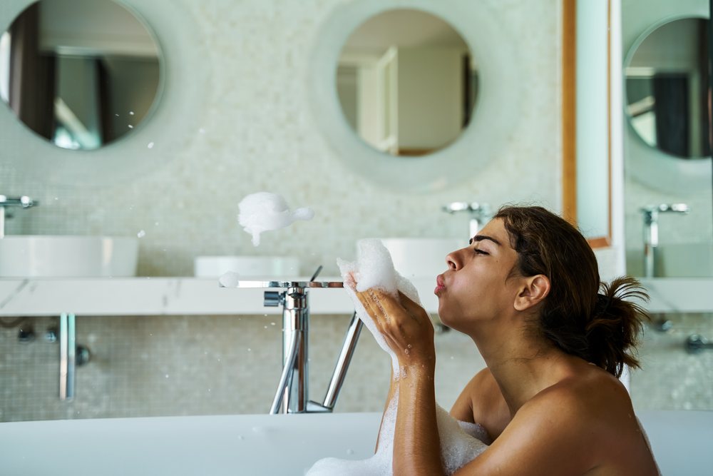 A white woman blowing foam bubbles in a bathtub in a modern bathroom