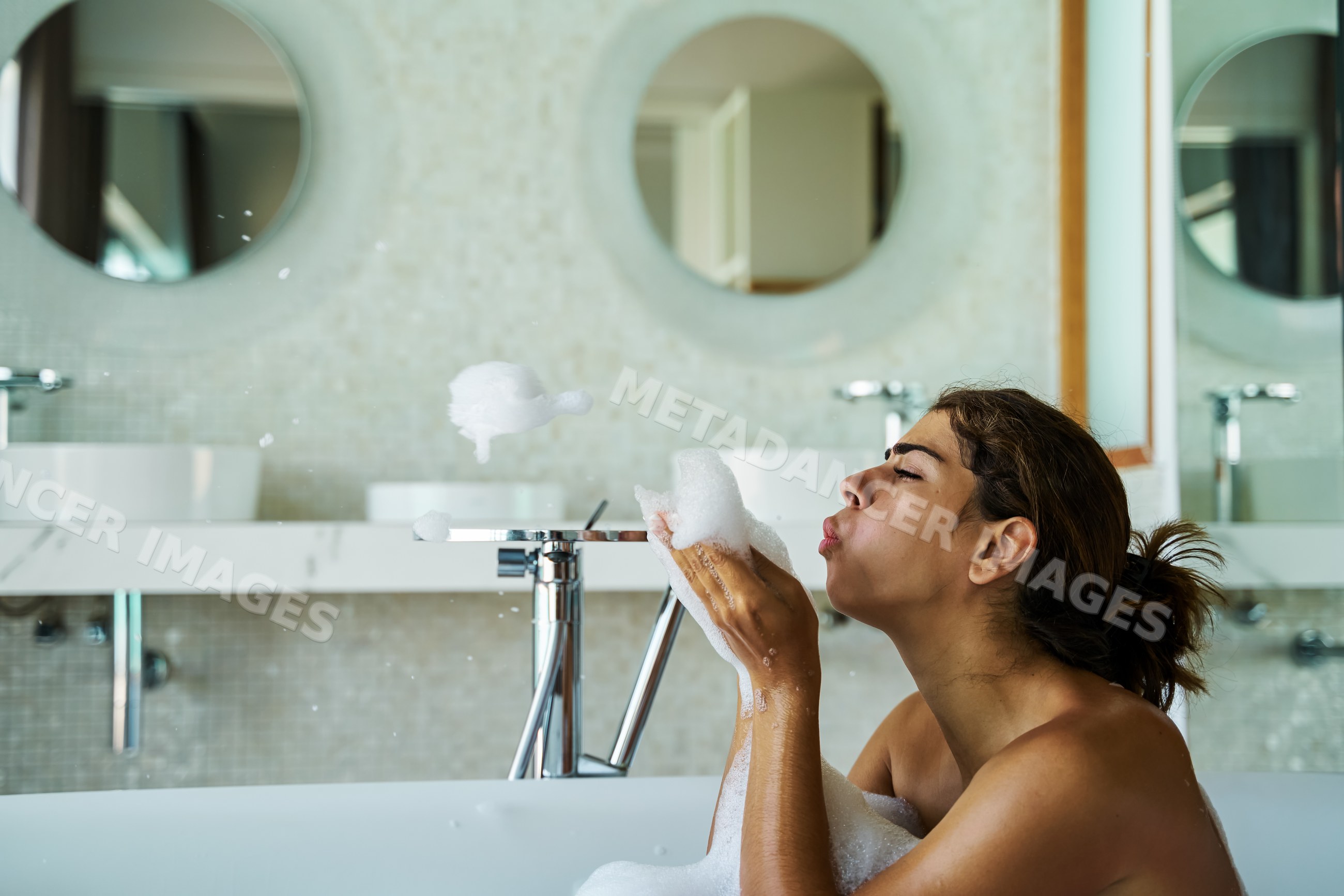 A white woman blowing foam bubbles in a bathtub in a modern bathroom