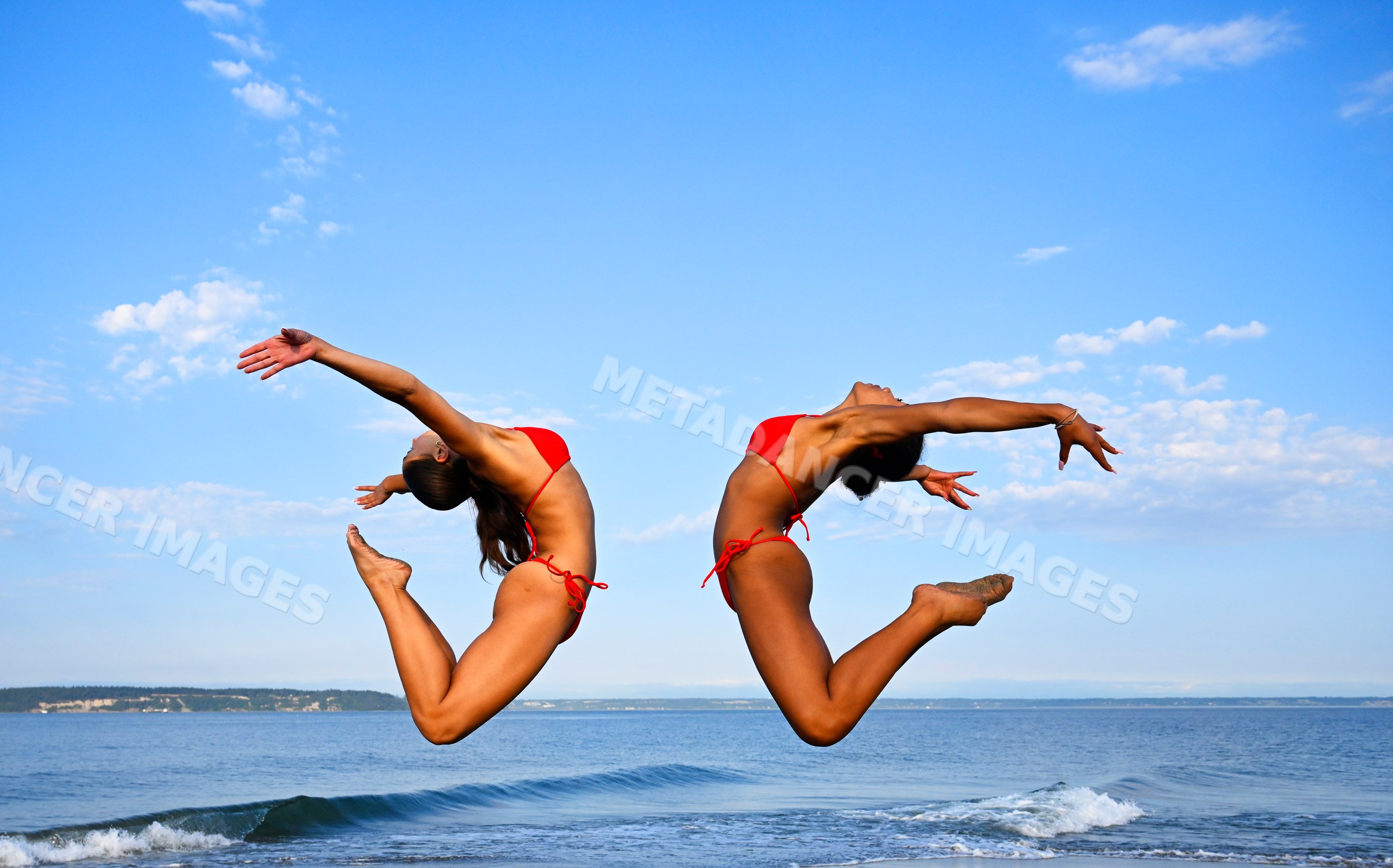 Two women perform synchronized partner yoga jumps on a beach