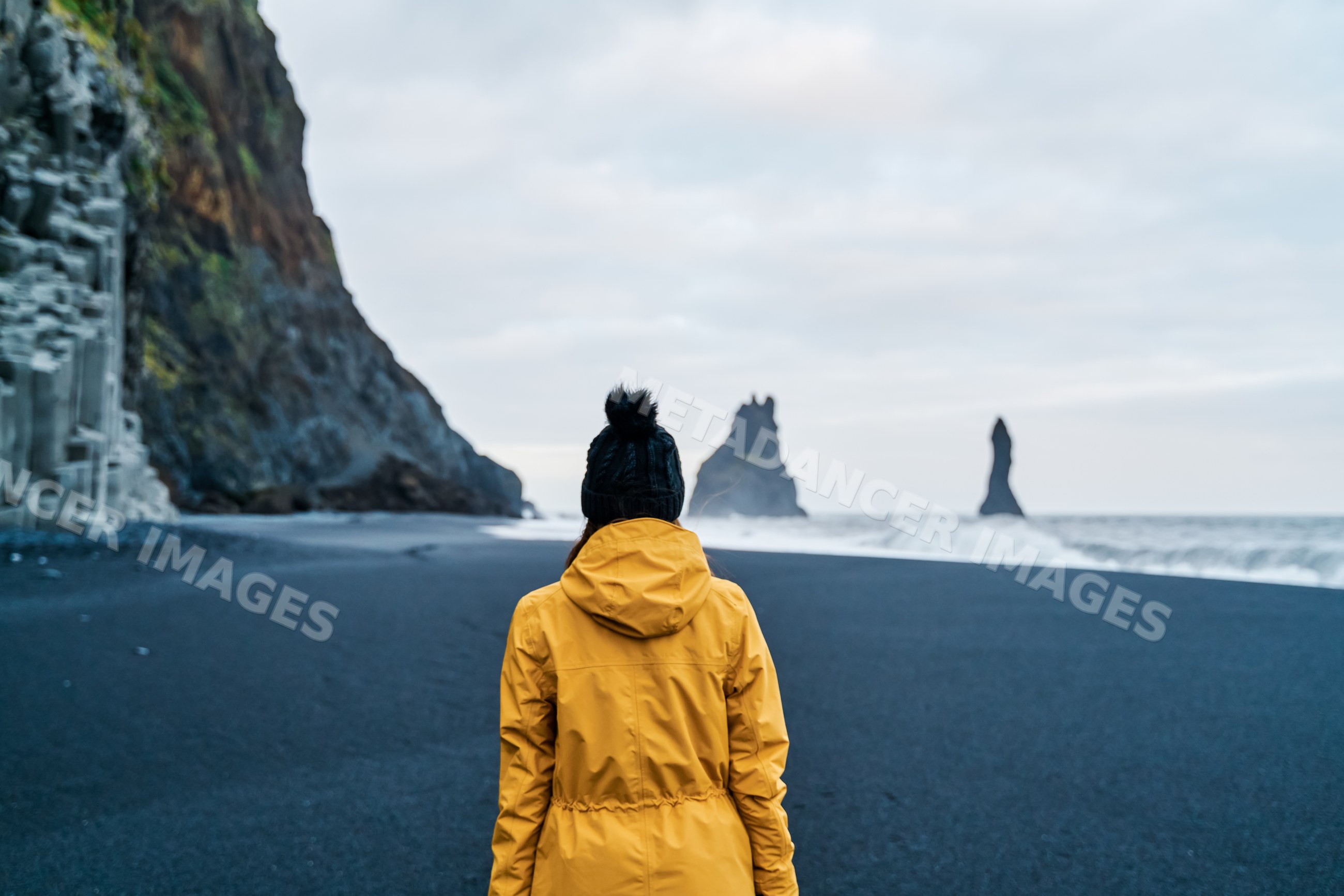 A white person in a yellow jacket walks on a black sand beach