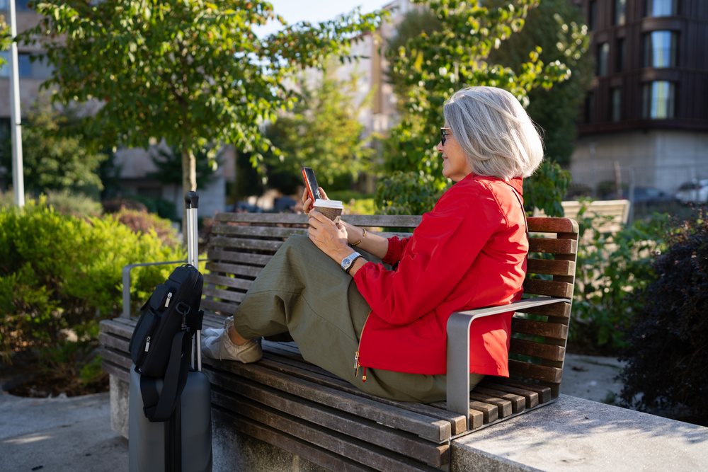 White woman sitting on a park bench using smartphone with coffee