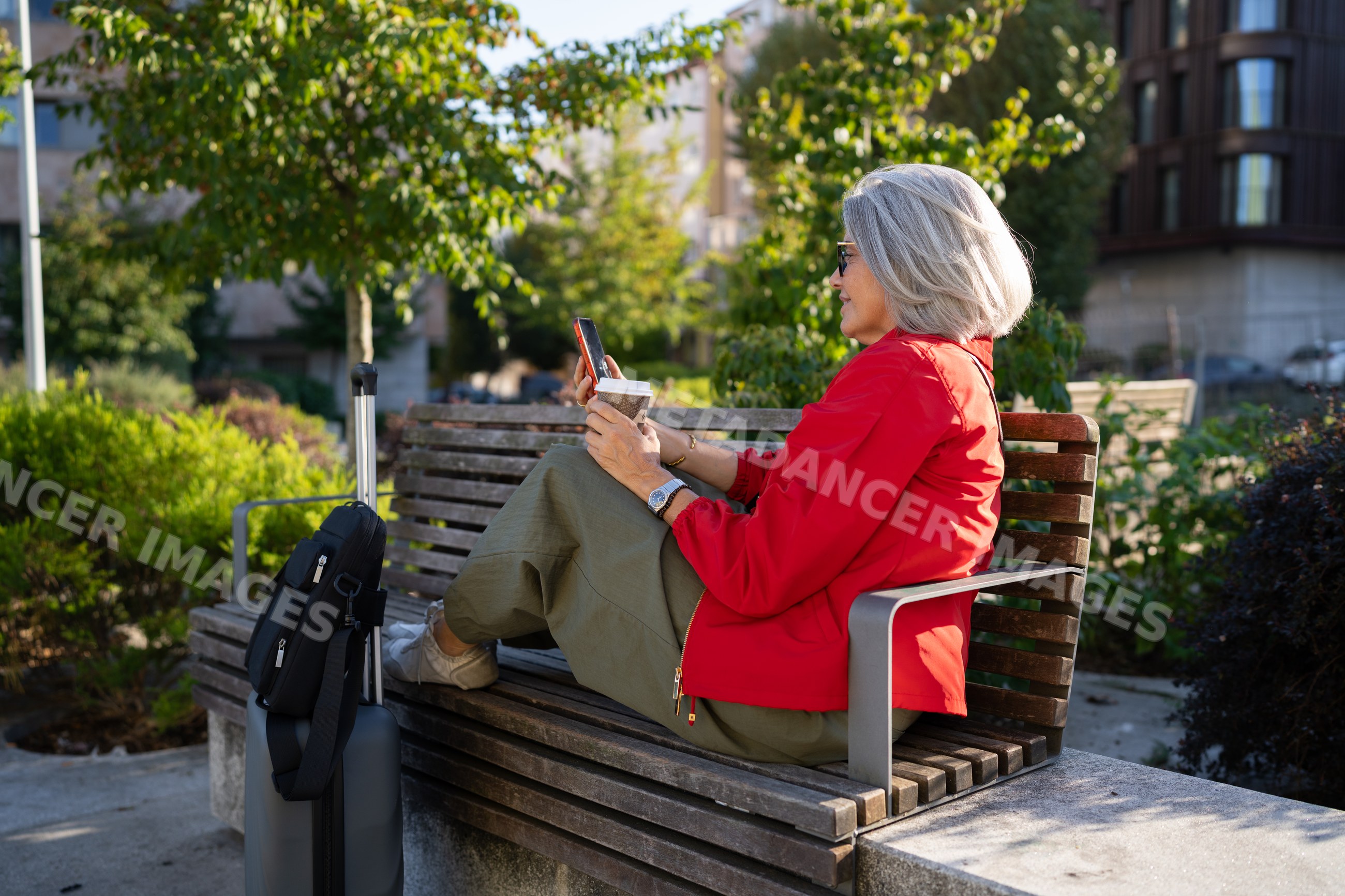 White woman sitting on a park bench using smartphone with coffee